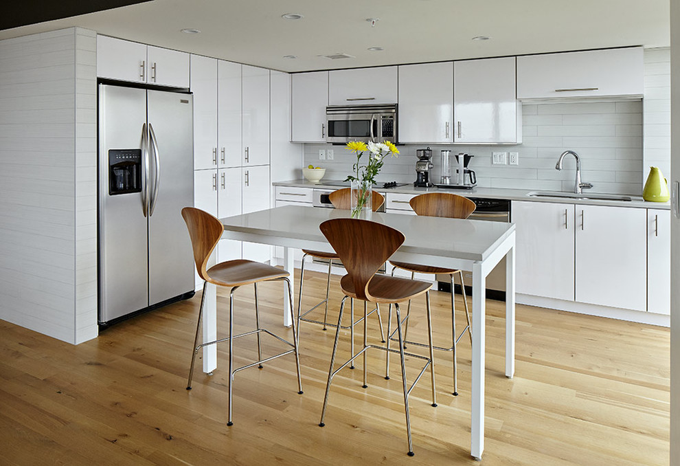 Exempel på ett modernt kök, med en undermonterad diskho, släta luckor, vita skåp, vitt stänkskydd, rostfria vitvaror, ljust trägolv och en köksö