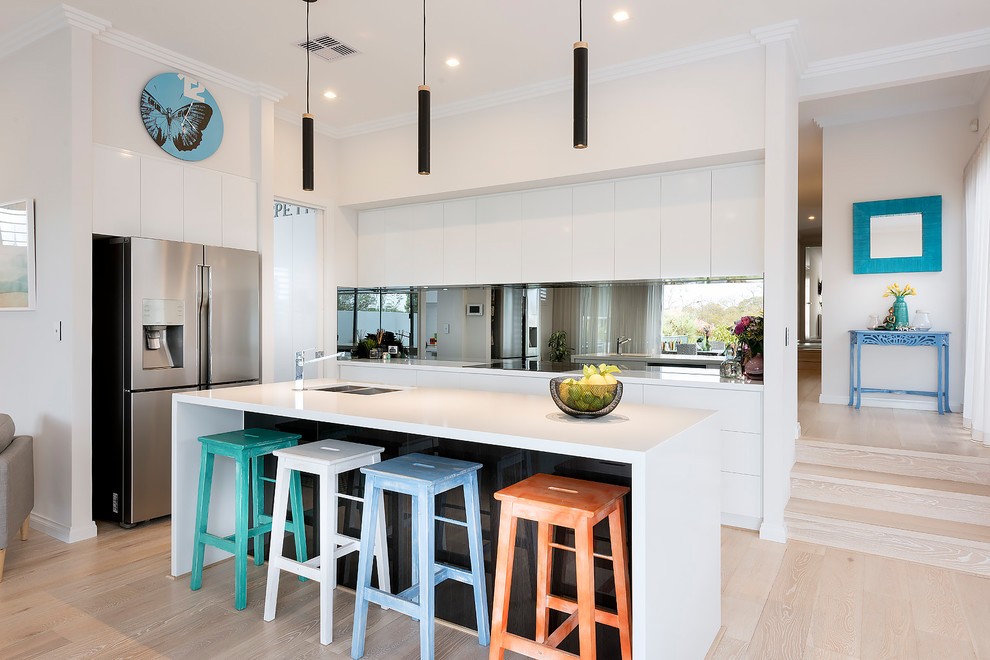 Modern inredning av ett kök, med släta luckor, vita skåp, spegel som stänkskydd, rostfria vitvaror, ljust trägolv, en köksö och beiget golv