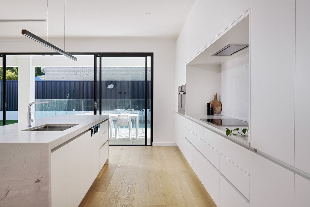 This is an example of a large modern galley kitchen/diner in Perth with a double-bowl sink, flat-panel cabinets, white cabinets, marble worktops, white splashback, marble splashback, black appliances, light hardwood flooring, an island, yellow floors and white worktops.