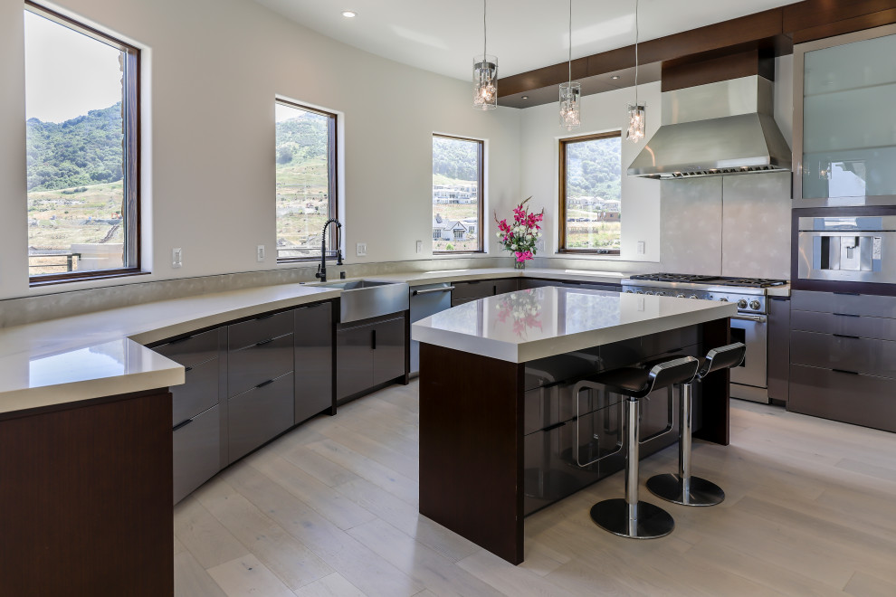 Trendy u-shaped light wood floor and beige floor kitchen photo in San Francisco with a farmhouse sink, flat-panel cabinets, brown cabinets, gray backsplash, stainless steel appliances, an island and beige countertops