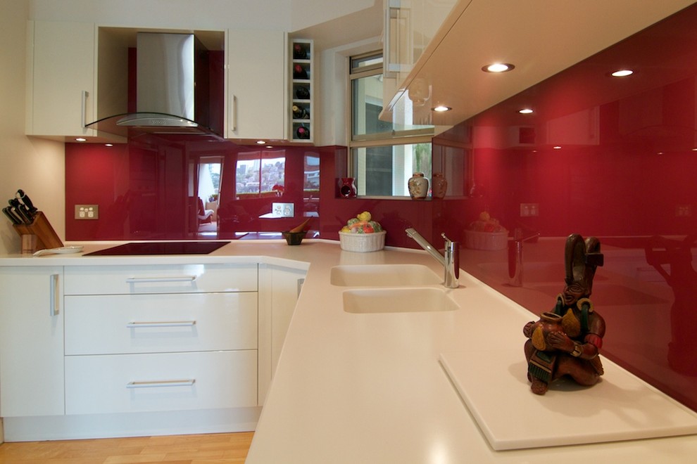 Small modern u-shaped enclosed kitchen in Sydney with an integrated sink, flat-panel cabinets, white cabinets, composite countertops, red splashback, glass sheet splashback, stainless steel appliances, light hardwood flooring and no island.