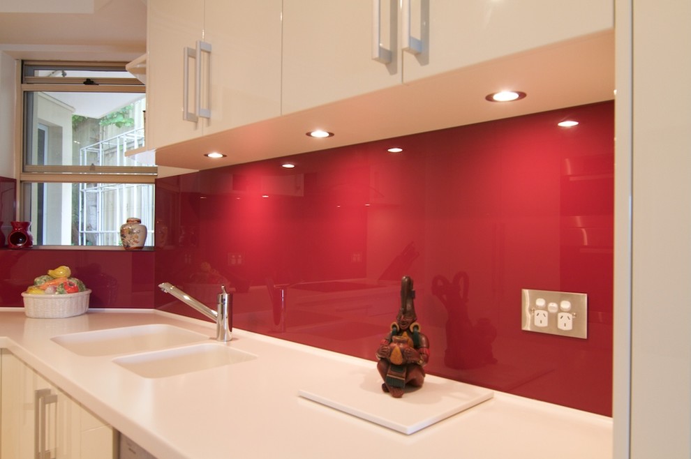 Example of a small minimalist u-shaped light wood floor enclosed kitchen design in Sydney with an integrated sink, flat-panel cabinets, white cabinets, solid surface countertops, red backsplash, glass sheet backsplash, stainless steel appliances and no island