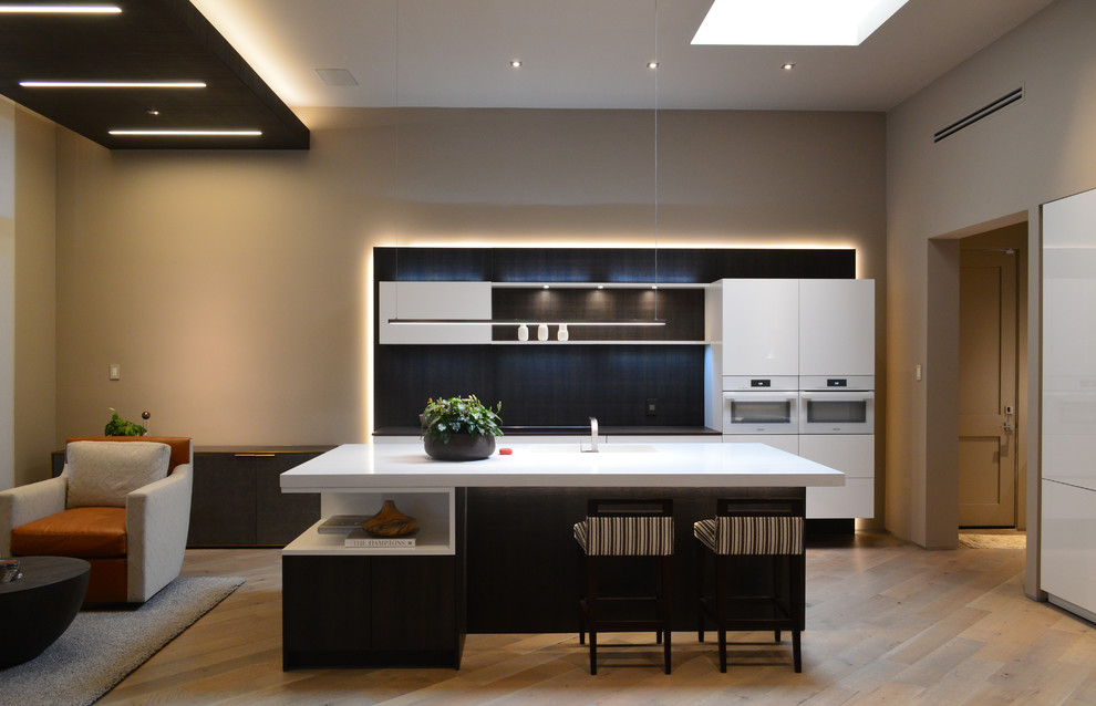 Свежая идея для дизайна: параллельная кухня-гостиная среднего размера в стиле модернизм с одинарной мойкой, белыми фасадами, черным фартуком, белой техникой, светлым паркетным полом, островом, коричневым полом и плоскими фасадами - отличное фото интерьера