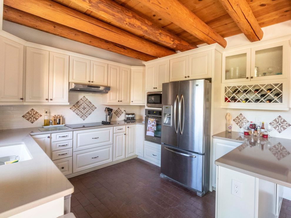 Example of a mid-sized transitional l-shaped brick floor and brown floor eat-in kitchen design in Albuquerque with an undermount sink, raised-panel cabinets, white cabinets, quartz countertops, white backsplash, porcelain backsplash, stainless steel appliances, a peninsula and gray countertops