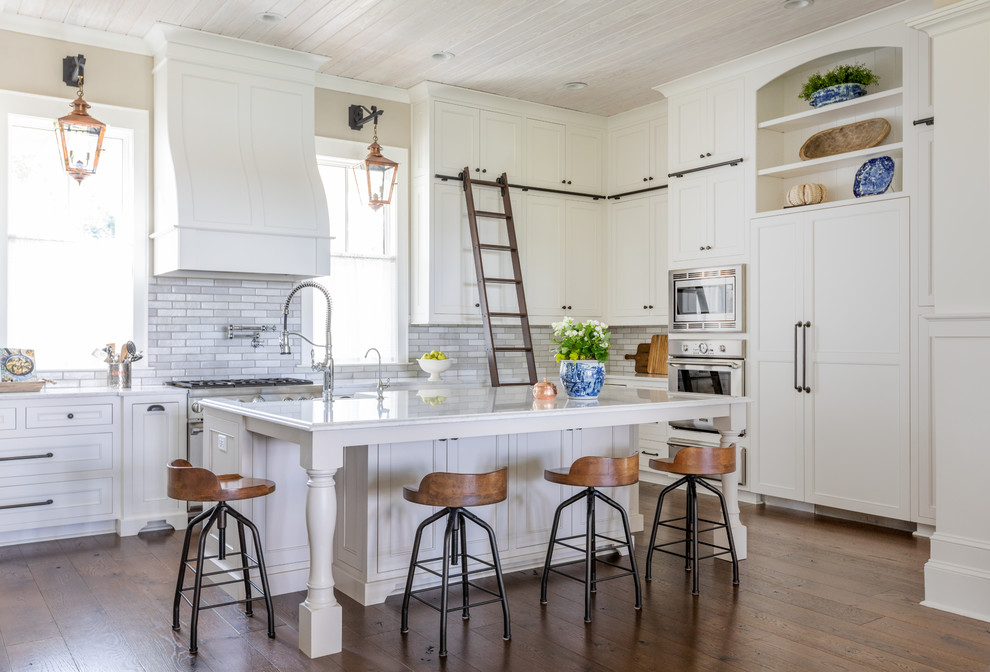 Lantlig inredning av ett vit vitt l-kök, med en rustik diskho, rostfria vitvaror, bänkskiva i kvarts, luckor med infälld panel, vita skåp, grått stänkskydd, mellanmörkt trägolv, en köksö och brunt golv