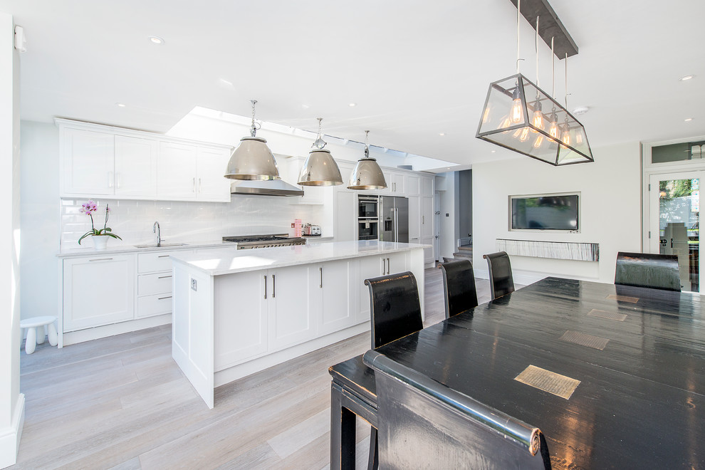 Inspiration for a large contemporary single-wall kitchen/diner in London with a built-in sink, recessed-panel cabinets, white cabinets, marble worktops, white splashback, metro tiled splashback, stainless steel appliances, light hardwood flooring and an island.