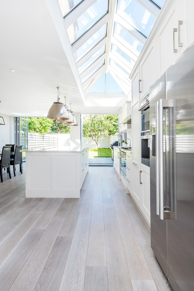 Zweizeilige Klassische Wohnküche mit weißen Schränken, Schrankfronten im Shaker-Stil, Küchengeräten aus Edelstahl, hellem Holzboden und Kücheninsel in London