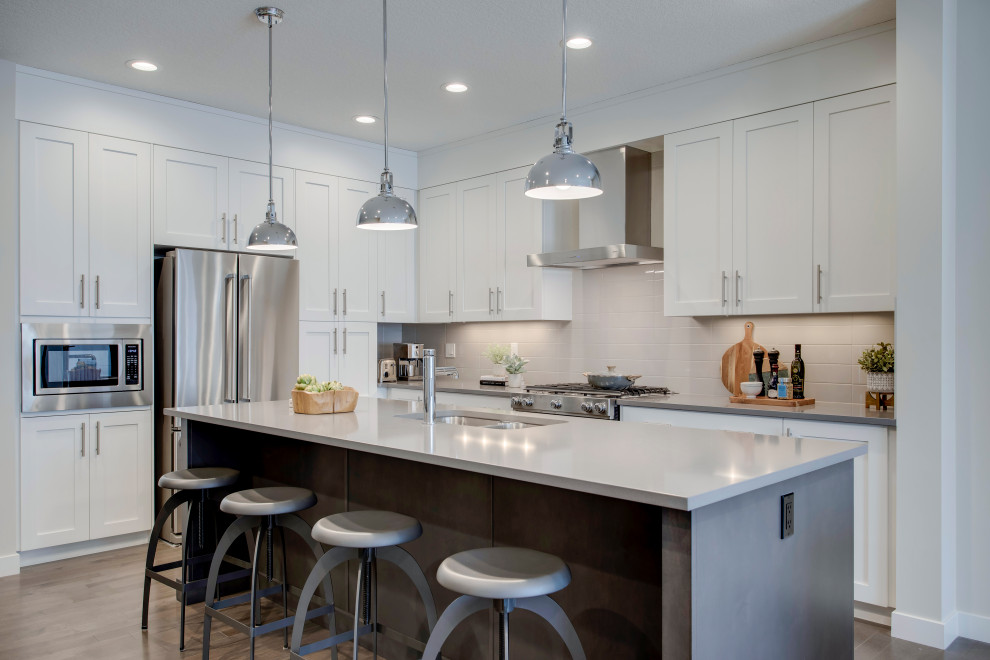Large classic l-shaped kitchen in Calgary with shaker cabinets, white cabinets, quartz worktops, white splashback, stainless steel appliances, medium hardwood flooring, an island, grey floors and grey worktops.