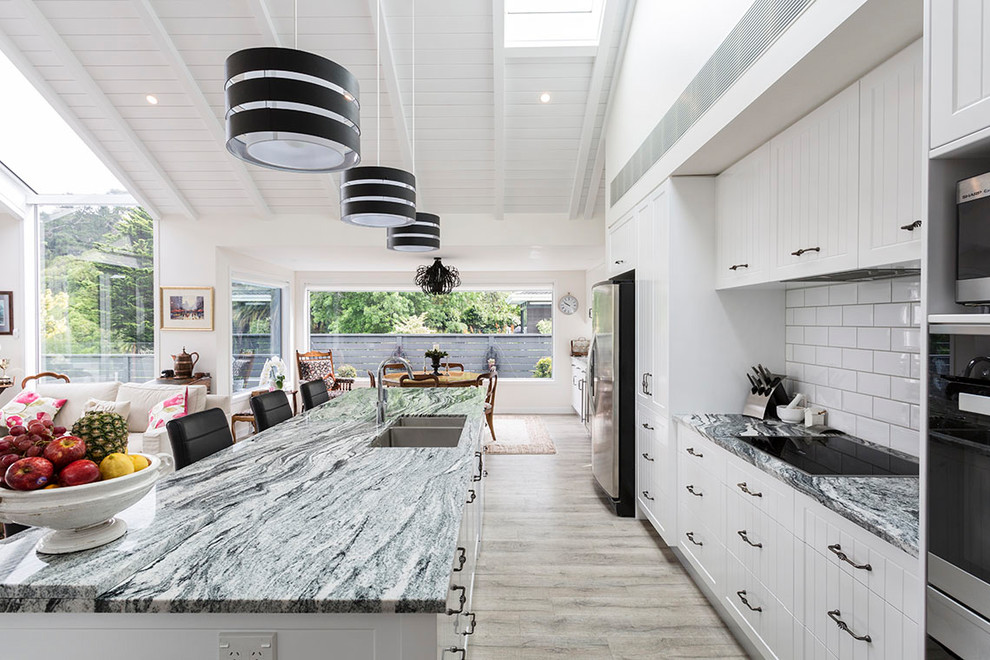 This is an example of a large farmhouse galley open plan kitchen in Other with a submerged sink, recessed-panel cabinets, white cabinets, granite worktops, white splashback, ceramic splashback, black appliances, light hardwood flooring, an island and grey floors.