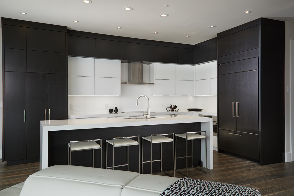 Photo of a medium sized contemporary l-shaped open plan kitchen in Toronto with a submerged sink, flat-panel cabinets, black cabinets, engineered stone countertops, white splashback, stone slab splashback, integrated appliances, medium hardwood flooring, an island and brown floors.