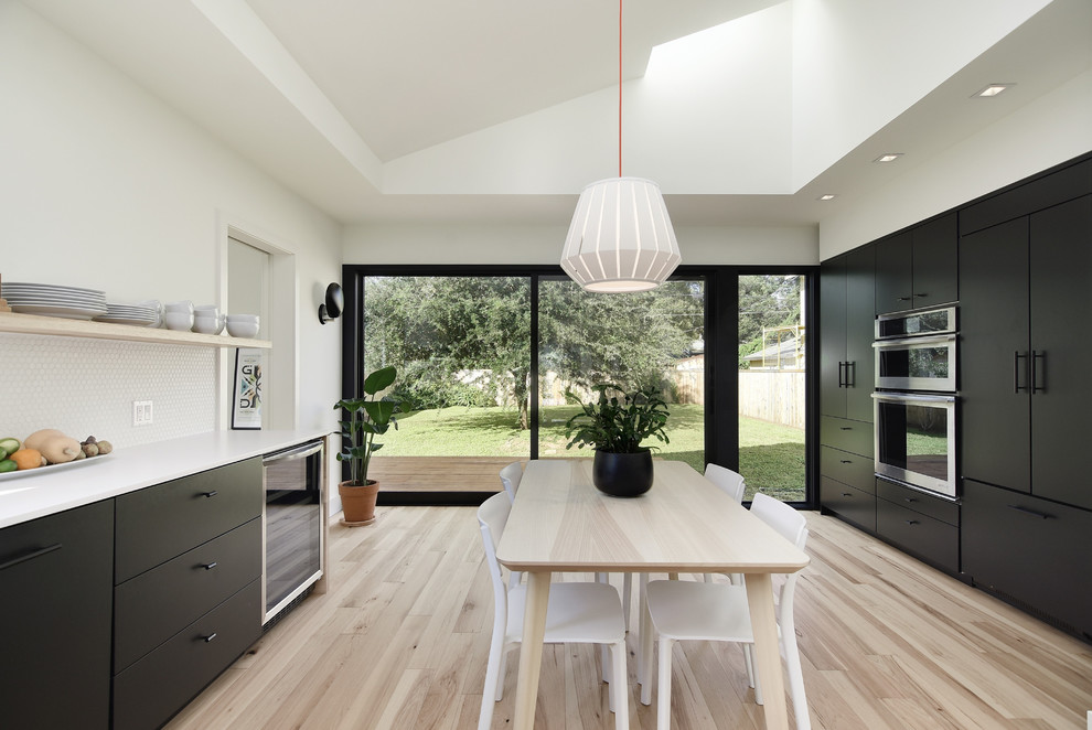 Example of a danish light wood floor and beige floor eat-in kitchen design in Austin with flat-panel cabinets, black cabinets, white backsplash, mosaic tile backsplash, stainless steel appliances and white countertops