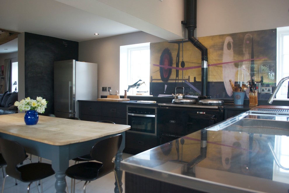 Photo of a medium sized modern l-shaped kitchen/diner in Wiltshire with an integrated sink, flat-panel cabinets, black cabinets, stainless steel worktops, multi-coloured splashback, glass sheet splashback, black appliances, concrete flooring and a breakfast bar.