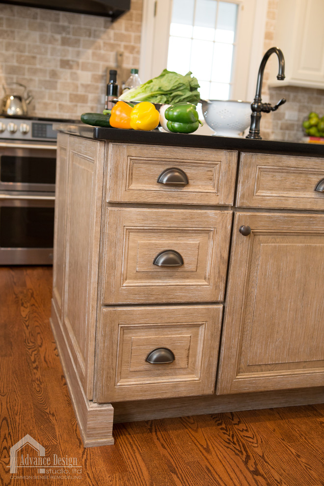 Mid-sized cottage u-shaped medium tone wood floor and brown floor eat-in kitchen photo in Chicago with a farmhouse sink, raised-panel cabinets, white cabinets, quartz countertops, beige backsplash, stone tile backsplash, stainless steel appliances, an island and black countertops