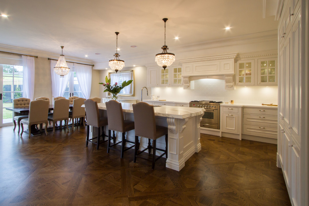 Klassisk inredning av ett stort vit vitt kök, med en undermonterad diskho, luckor med infälld panel, vitt stänkskydd, stänkskydd i keramik, mellanmörkt trägolv, en köksö, vita skåp, marmorbänkskiva, rostfria vitvaror och brunt golv