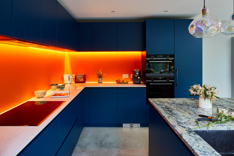Cette photo montre une grande cuisine américaine tendance en L avec un placard à porte plane, des portes de placard bleues, un plan de travail en quartz, une crédence orange, une crédence en feuille de verre, un électroménager en acier inoxydable, sol en béton ciré, îlot et un sol gris.