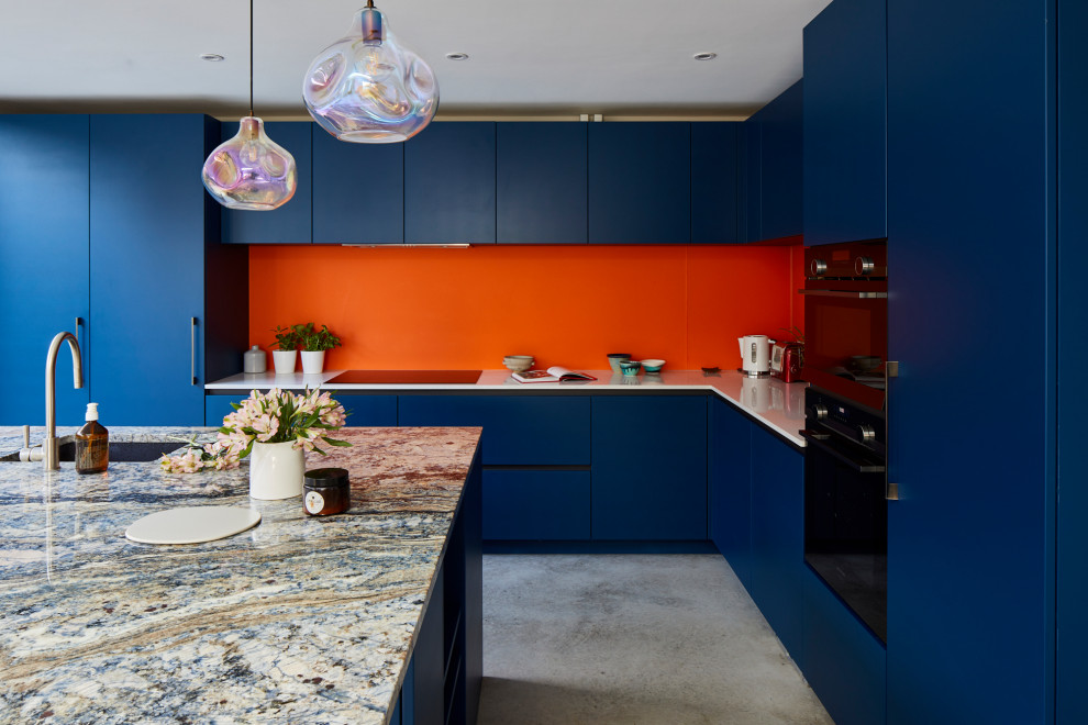 Große Moderne Wohnküche in L-Form mit flächenbündigen Schrankfronten, blauen Schränken, Quarzit-Arbeitsplatte, Küchenrückwand in Orange, Glasrückwand, Küchengeräten aus Edelstahl, Betonboden, Kücheninsel und grauem Boden in London