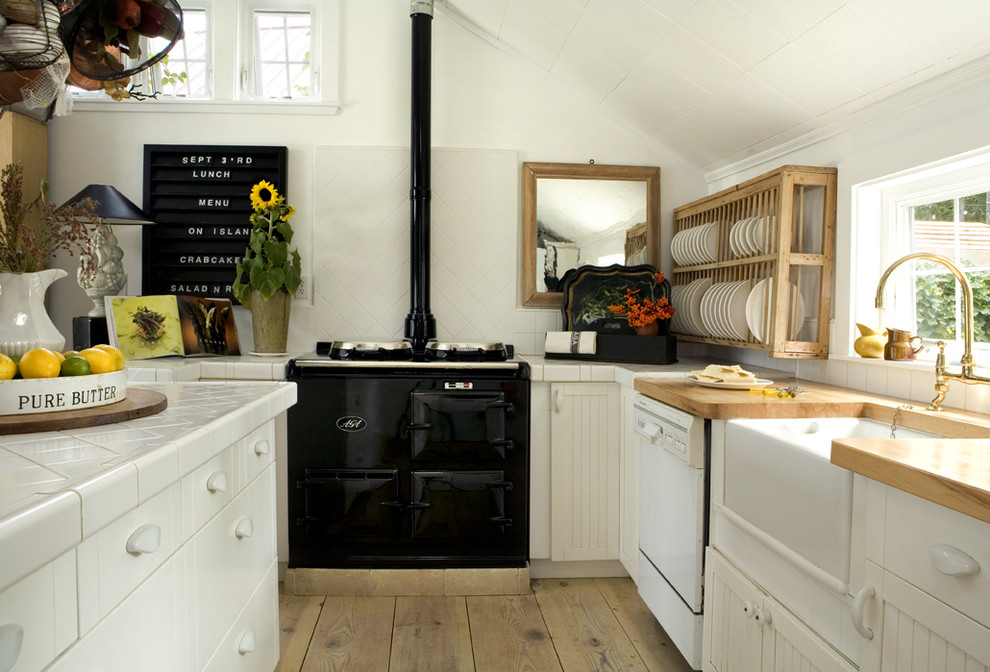 ポートランドにある北欧スタイルのおしゃれなキッチン (エプロンフロントシンク、タイルカウンター、オープンシェルフ、白いキャビネット、黒い調理設備、白いキッチンパネル) の写真