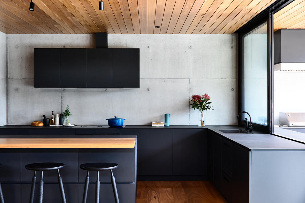 Contemporary l-shaped kitchen in Melbourne with a built-in sink, flat-panel cabinets, black cabinets, grey splashback, cement tile splashback, black appliances, an island, black worktops, medium hardwood flooring and brown floors.