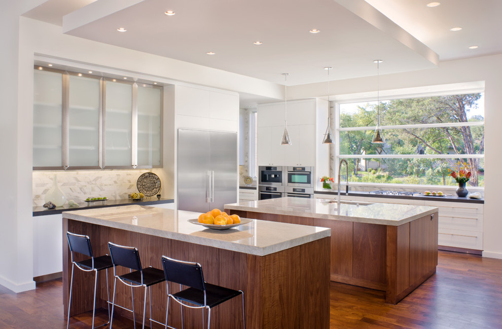 Bild på ett funkis kök, med luckor med glaspanel, skåp i rostfritt stål, vitt stänkskydd, stänkskydd i stenkakel, rostfria vitvaror, mörkt trägolv och flera köksöar