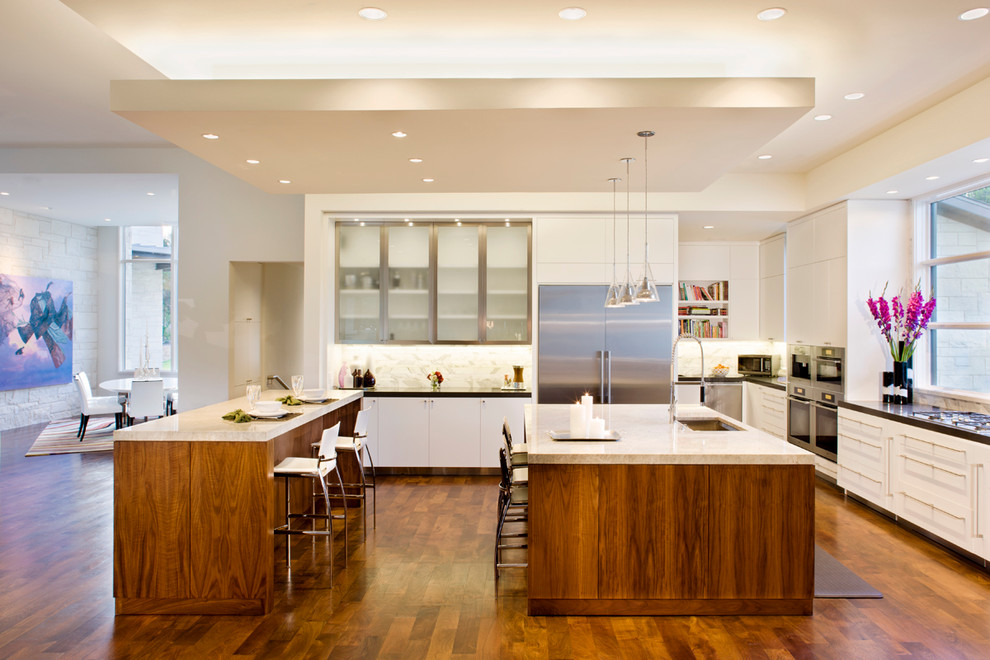 Example of a trendy galley dark wood floor open concept kitchen design in Austin with white cabinets, flat-panel cabinets, white backsplash, stone tile backsplash, stainless steel appliances, two islands and an undermount sink