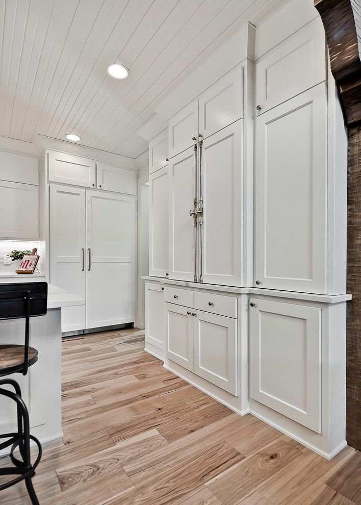 Amerikansk inredning av ett stort vit vitt kök med öppen planlösning, med en rustik diskho, luckor med upphöjd panel, vita skåp, bänkskiva i kvartsit, vitt stänkskydd, stänkskydd i stenkakel, rostfria vitvaror, ljust trägolv, en köksö och brunt golv