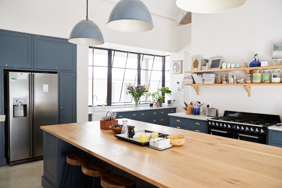 Modern inredning av ett mellanstort beige beige kök, med en rustik diskho, luckor med profilerade fronter, blå skåp, träbänkskiva, vitt stänkskydd, fönster som stänkskydd, svarta vitvaror, klinkergolv i porslin, en köksö och beiget golv
