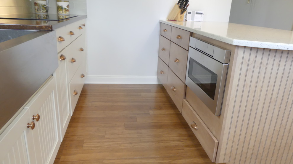 This is an example of a medium sized country l-shaped enclosed kitchen in New York with a belfast sink, beaded cabinets, white cabinets, engineered stone countertops, green splashback, glass tiled splashback, stainless steel appliances, medium hardwood flooring, a breakfast bar and brown floors.