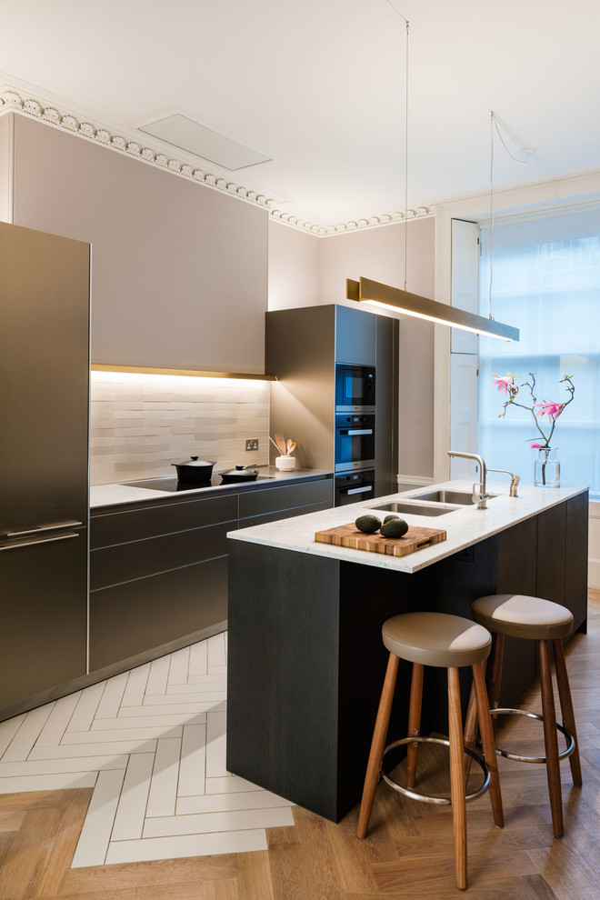 This is an example of a large contemporary galley kitchen in London with flat-panel cabinets, brown cabinets, marble worktops, beige splashback, ceramic splashback, black appliances, light hardwood flooring, an island and a submerged sink.