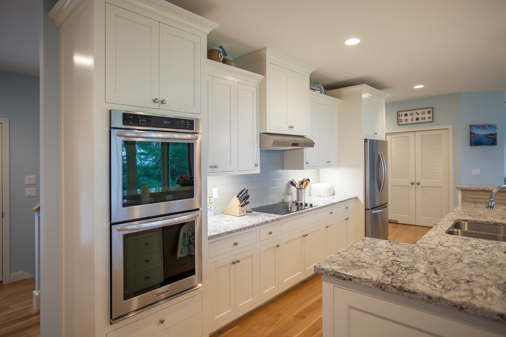 Photo of a coastal single-wall open plan kitchen in Grand Rapids with shaker cabinets, white cabinets, granite worktops, white splashback, porcelain splashback, stainless steel appliances, light hardwood flooring and an island.