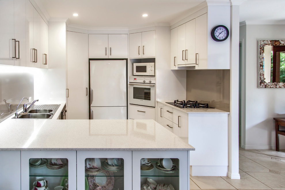 Bild på ett stort funkis kök, med en nedsänkt diskho, släta luckor, vita skåp, bänkskiva i kvarts, brunt stänkskydd, glaspanel som stänkskydd, vita vitvaror, klinkergolv i keramik och en köksö