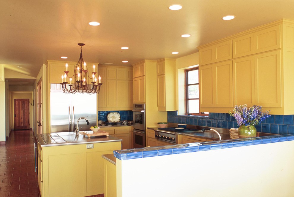 サンルイスオビスポにある地中海スタイルのおしゃれなコの字型キッチン (シルバーの調理設備、ステンレスカウンター、シェーカースタイル扉のキャビネット、黄色いキャビネット、青いキッチンパネル) の写真