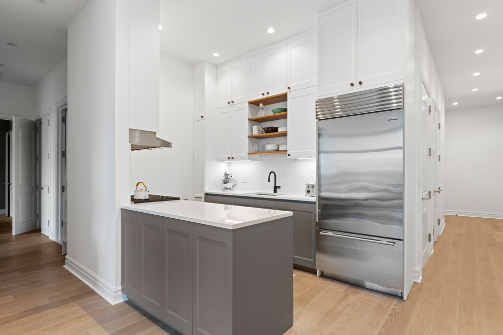 Photo of a medium sized classic galley enclosed kitchen in New York with a submerged sink, recessed-panel cabinets, grey cabinets, engineered stone countertops, white splashback, stone slab splashback, stainless steel appliances, medium hardwood flooring, a breakfast bar, brown floors and white worktops.