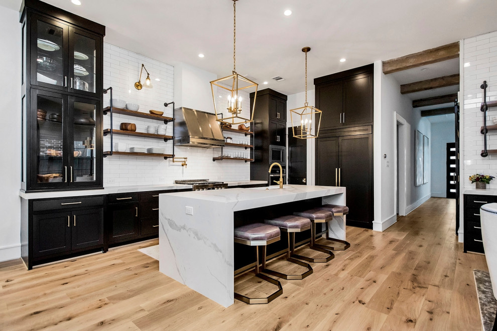 Example of a transitional l-shaped medium tone wood floor and brown floor kitchen design in Houston with a farmhouse sink, shaker cabinets, black cabinets, white backsplash, subway tile backsplash, an island and white countertops