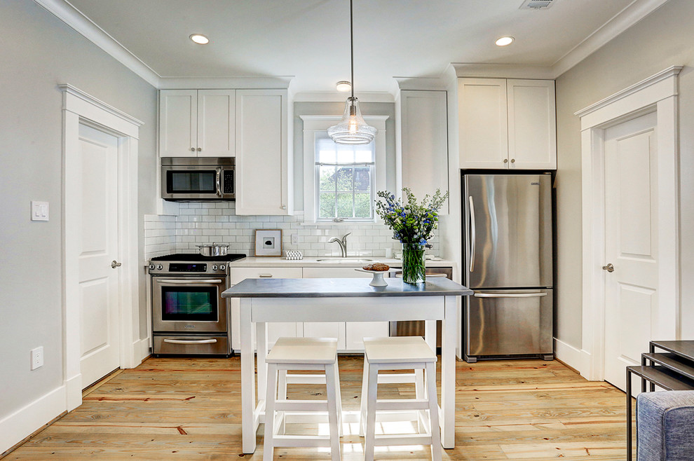 Klassisk inredning av ett litet linjärt kök med öppen planlösning, med en undermonterad diskho, skåp i shakerstil, vita skåp, vitt stänkskydd, stänkskydd i tunnelbanekakel, rostfria vitvaror, ljust trägolv, en köksö och brunt golv