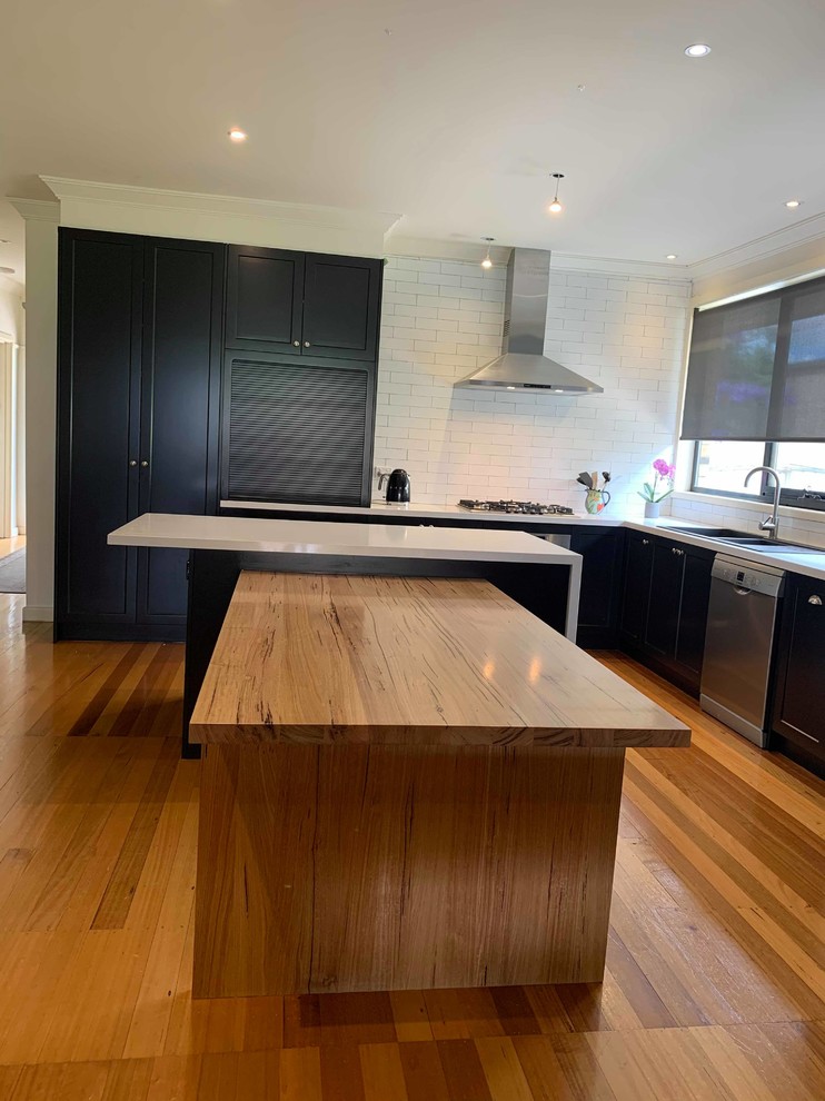 This is an example of a large contemporary l-shaped open plan kitchen in Melbourne with shaker cabinets, an island, a double-bowl sink, black cabinets, granite worktops, white splashback, porcelain splashback, stainless steel appliances, medium hardwood flooring and white worktops.