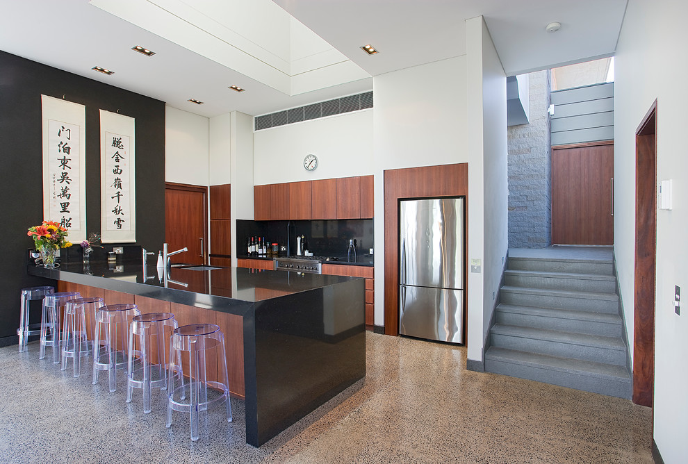 This is an example of a large world-inspired galley kitchen in Sydney with a submerged sink, flat-panel cabinets, medium wood cabinets, engineered stone countertops, black splashback, stone slab splashback, stainless steel appliances, concrete flooring, grey floors, black worktops and a breakfast bar.