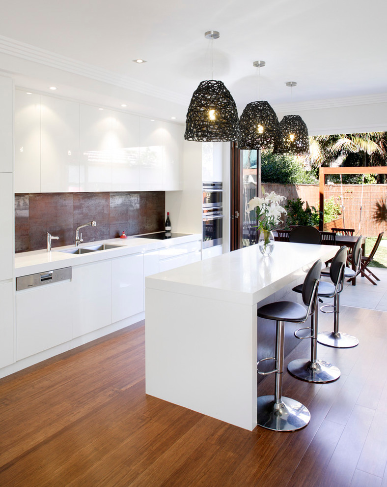 Inspiration for a large contemporary galley open plan kitchen in Sydney with a submerged sink, flat-panel cabinets, white cabinets, engineered stone countertops, metallic splashback, stainless steel appliances, an island, brown floors, white worktops, porcelain splashback and medium hardwood flooring.