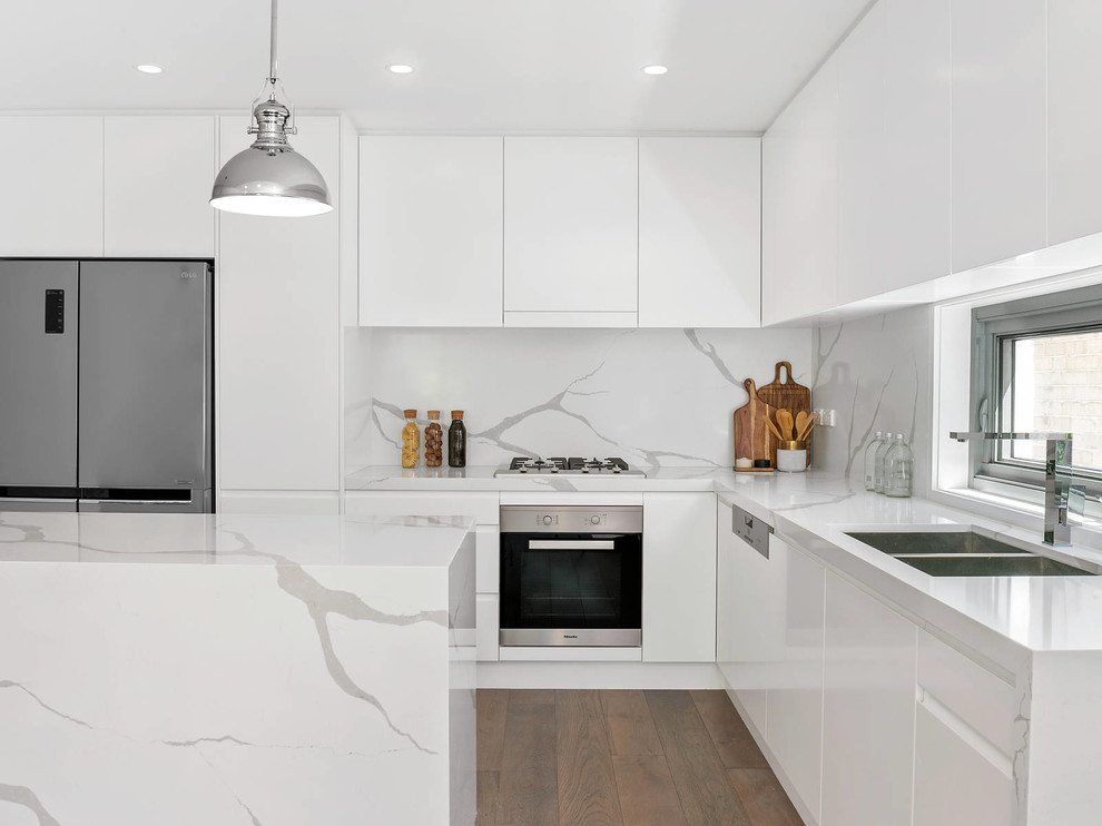 Contemporary l-shaped kitchen/diner in Sydney with a double-bowl sink, white cabinets, stainless steel appliances, an island, brown floors, flat-panel cabinets, white splashback, stone slab splashback, dark hardwood flooring and white worktops.
