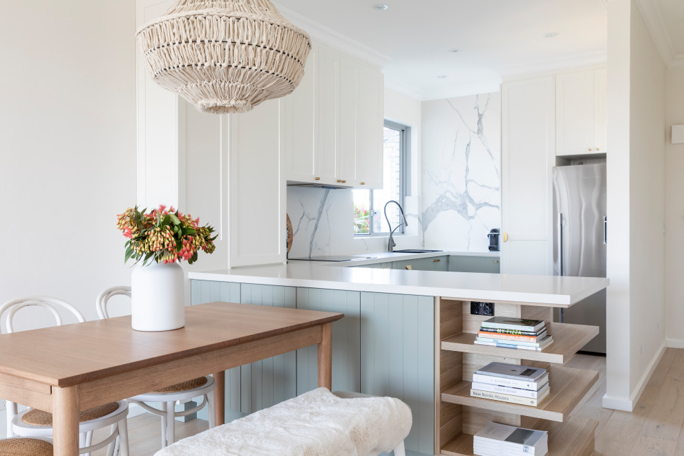 Photo of a small nautical u-shaped kitchen/diner in Sydney with a submerged sink, shaker cabinets, white cabinets, engineered stone countertops, white splashback, marble splashback, stainless steel appliances, light hardwood flooring, white worktops, a breakfast bar and beige floors.