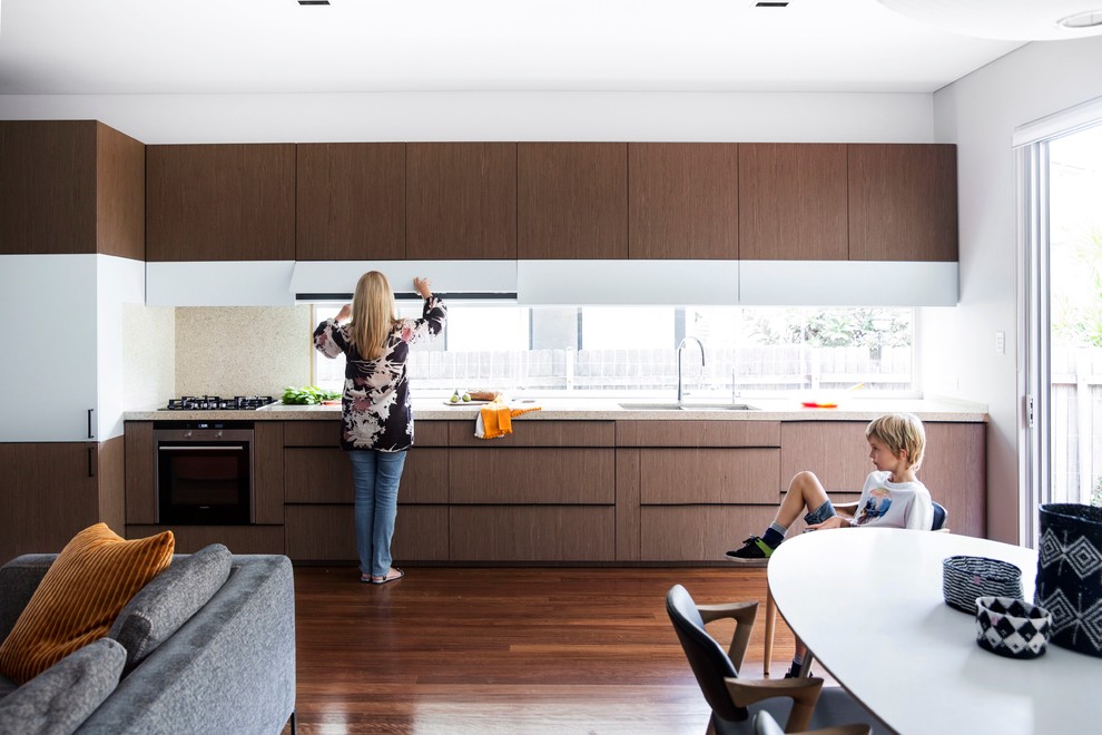 Modelo de cocina lineal contemporánea de tamaño medio abierta sin isla con fregadero bajoencimera, armarios con paneles lisos, puertas de armario de madera oscura, encimera de cuarcita, electrodomésticos de acero inoxidable y suelo de madera en tonos medios