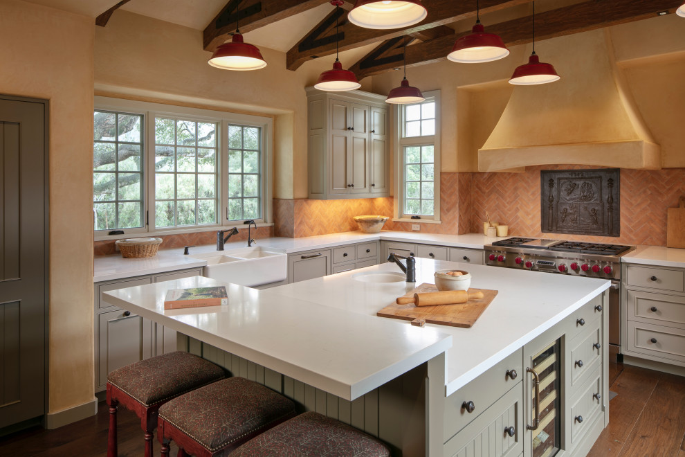 Example of a tuscan l-shaped medium tone wood floor and vaulted ceiling kitchen design in Other with a farmhouse sink, beaded inset cabinets, gray cabinets, orange backsplash, stainless steel appliances, an island and white countertops