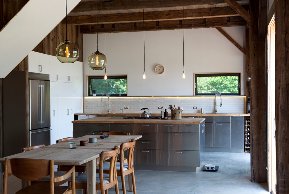 Example of a mid-sized mountain style concrete floor and gray floor eat-in kitchen design in New York with subway tile backsplash, stainless steel cabinets, wood countertops, white backsplash, flat-panel cabinets, stainless steel appliances, an undermount sink and an island