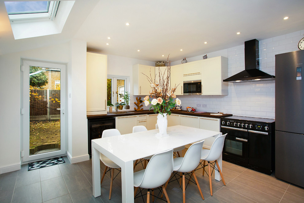 Contemporary l-shaped kitchen/diner in London with a built-in sink, flat-panel cabinets, white cabinets, wood worktops, white splashback, metro tiled splashback, black appliances, no island, beige floors and brown worktops.