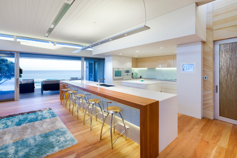 Moderne Küche in U-Form mit flächenbündigen Schrankfronten, weißen Schränken, weißen Elektrogeräten, braunem Holzboden und zwei Kücheninseln in Sonstige
