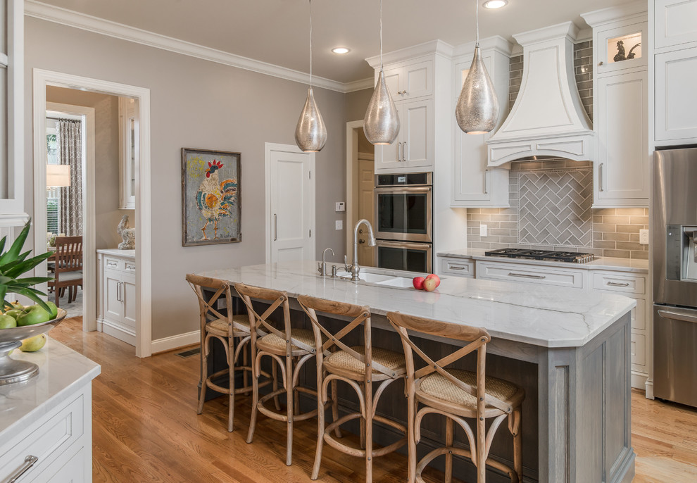 Example of a transitional galley medium tone wood floor and brown floor kitchen design in Nashville with white cabinets, gray backsplash, subway tile backsplash, stainless steel appliances, an island, an undermount sink, recessed-panel cabinets and white countertops