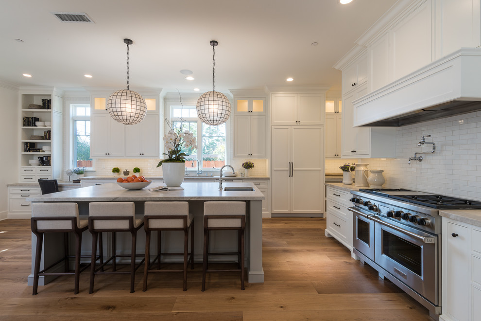 This is an example of a classic l-shaped kitchen in Los Angeles with a submerged sink, shaker cabinets, white cabinets, white splashback, stainless steel appliances, medium hardwood flooring, an island and brown floors.