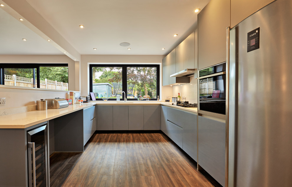 This is an example of a contemporary u-shaped kitchen in Essex with flat-panel cabinets, grey cabinets, beige splashback, stainless steel appliances, dark hardwood flooring, a breakfast bar, brown floors and beige worktops.