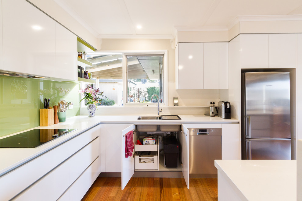 Modern inredning av ett mellanstort vit vitt kök, med en dubbel diskho, släta luckor, vita skåp, bänkskiva i kvarts, grönt stänkskydd, glaspanel som stänkskydd, rostfria vitvaror, mellanmörkt trägolv, en köksö och brunt golv