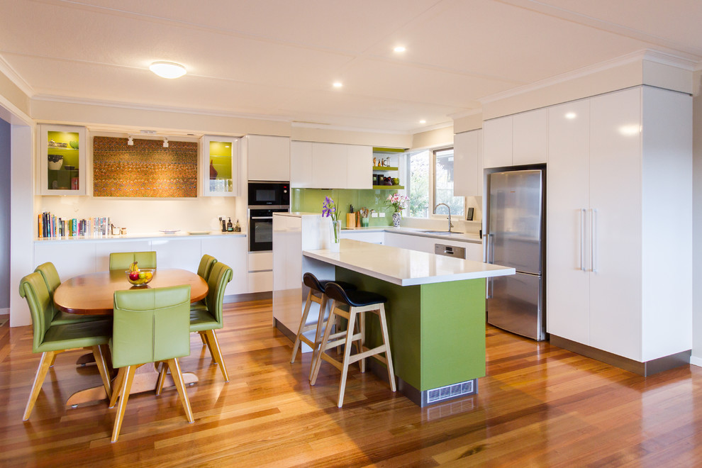 Eat-in kitchen - mid-sized contemporary l-shaped medium tone wood floor and brown floor eat-in kitchen idea in Melbourne with a double-bowl sink, flat-panel cabinets, white cabinets, quartz countertops, green backsplash, glass sheet backsplash, stainless steel appliances, an island and white countertops