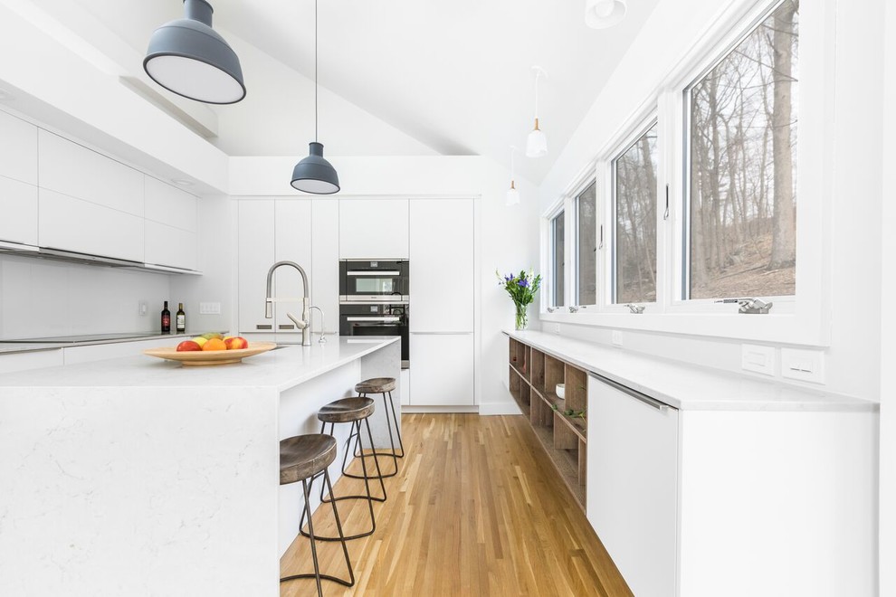 Mid-sized minimalist galley medium tone wood floor and brown floor eat-in kitchen photo in New York with a drop-in sink, white cabinets, quartz countertops, white backsplash, glass sheet backsplash, stainless steel appliances and an island